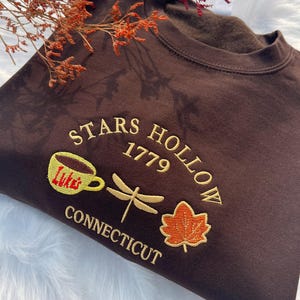 May include: Brown sweatshirt with embroidered text "Stars Hollow 1779 Connecticut" and images of a coffee cup, dragonfly, and maple leaf.
