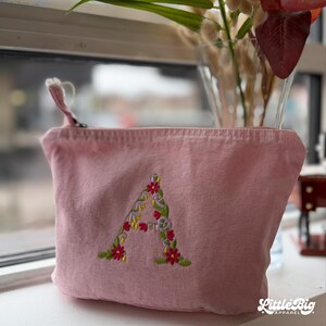 Puede incluir: Una bolsa con cremallera de lona rosa con una letra "A" floral bordada. La letra está llena de flores rojas, amarillas y moradas y hojas verdes. La bolsa tiene un pequeño lazo en la cremallera.