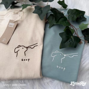 May include: Two sweatshirts, one ivory and one jade green, with embroidered dog boop designs. The ivory sweatshirt has brown embroidery, the jade green has white. The word "boop" is below the design. The tag reads "Organic Cotton Cotton BIO".