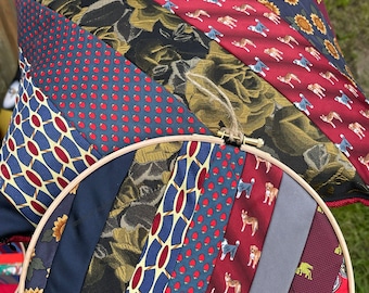 Può includere: Primo piano di un cerchio da ricamo circolare con un patchwork colorato di cravatte in tessuto. Il tessuto è disposto in strisce diagonali e presenta vari motivi, tra cui pois, disegni floreali e forme geometriche.