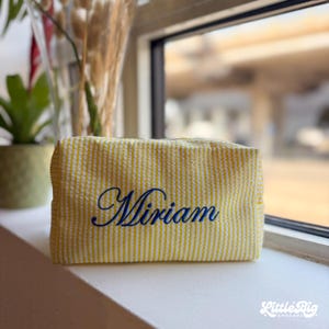 Puede incluir: Un estuche rectangular con cremallera hecho de tela seersucker amarilla y blanca con un patrón de rayas texturizado. El nombre Miriam está bordado en una fuente cursiva de color azul oscuro en el centro del panel frontal del estuche.