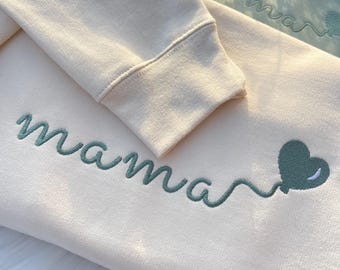 May include: A cream-colored sweatshirt featuring the embroidered word mama in sage green cursive script, ending in a sage green heart-shaped balloon. The sleeve is embroidered with the name Ellie in sage green above a small red heart.
