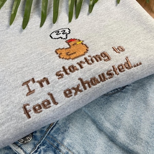 Puede incluir: Una sudadera gris claro con la frase bordada "I'm starting to feel exhausted..." en hilo marr&oacute;n. El dise&ntilde;o incluye un brazo pixelado con una burbuja de pensamiento. La sudadera se combina con una chaqueta vaquera.