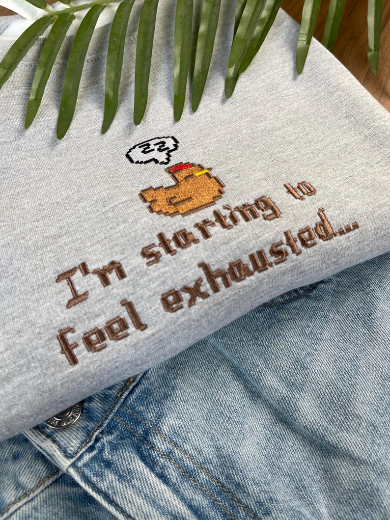 Puede incluir: Una sudadera gris claro con la frase bordada "I'm starting to feel exhausted..." en hilo marr&oacute;n. El dise&ntilde;o incluye un brazo pixelado con una burbuja de pensamiento. La sudadera se combina con una chaqueta vaquera.