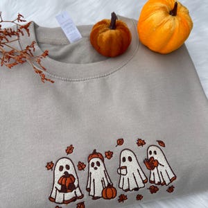 May include: A beige sweatshirt with an embroidered design of four cartoon ghosts holding pumpkins, a pumpkin, and a cup of coffee. The ghosts are surrounded by embroidered autumn leaves. Two small orange pumpkins sit on top of the sweatshirt.