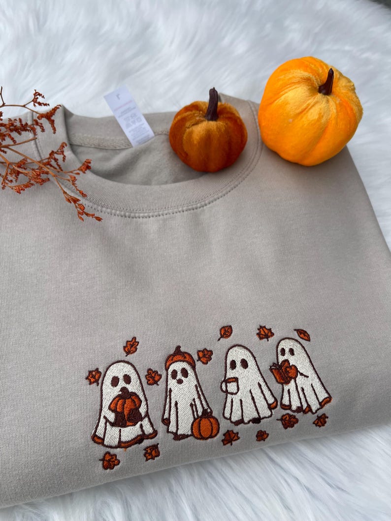 May include: A beige sweatshirt with an embroidered design of four cartoon ghosts holding pumpkins, a pumpkin, and a cup of coffee. The ghosts are surrounded by embroidered autumn leaves. Two small orange pumpkins sit on top of the sweatshirt.