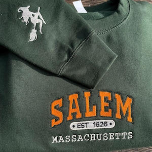 Peut inclure: Sweat-shirt vert fonc&eacute; avec le mot "SALEM" en lettres orange, bord&eacute;es de noir, et le texte "EST 1626" et "MASSACHUSETTS" en dessous. Une sorci&egrave;re blanche brod&eacute;e est sur la manche gauche.