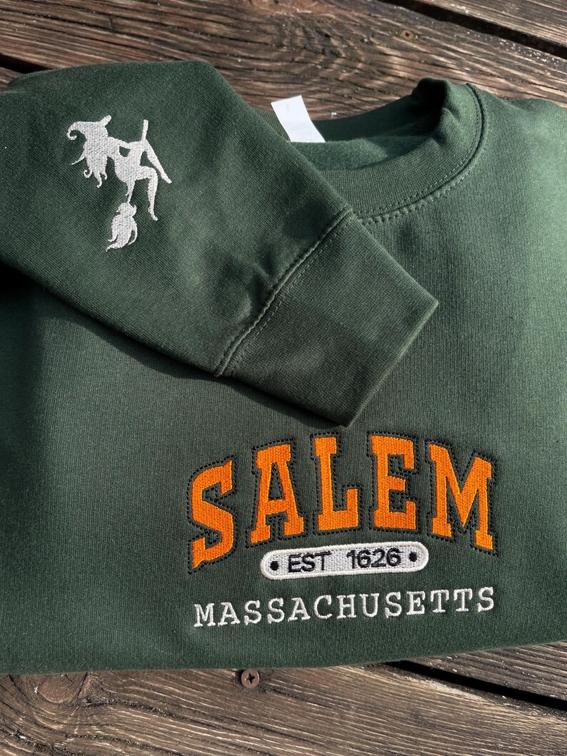 Peut inclure: Sweat-shirt vert fonc&eacute; avec le mot "SALEM" en lettres orange, bord&eacute;es de noir, et le texte "EST 1626" et "MASSACHUSETTS" en dessous. Une sorci&egrave;re blanche brod&eacute;e est sur la manche gauche.