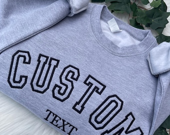 May include: A light grey crewneck sweatshirt featuring the word CUSTOM in large, dark navy blue block letters with a thin outline, positioned above the word TEXT in a smaller, matching font. The garment has long sleeves and a ribbed collar.