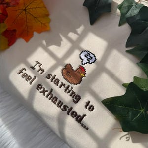 Puede incluir: Sudadera color crema con texto bordado y un gr&aacute;fico de pollo pixelado. El texto dice "I'm starting to feel exhausted..." con una burbuja de pensamiento que muestra s&iacute;mbolos "zz". Hojas verdes y follaje oto&ntilde;al rodean la sudadera.