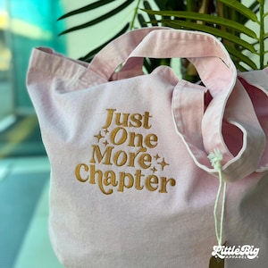 Op de afbeelding: Een lichtroze canvas draagtas met de geborduurde gouden tekst Just One More Chapter en kleine steraccenten. De tas heeft twee stoffen handvatten en een wit label aan de zijkant met de merknaam Little Big Apparel erop vermeld.