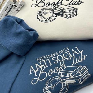 Può includere: Due felpe, una color crema e una blu navy, ricamate con la scritta "MEMBERS ONLY ANTI SOCIAL Book club". Ogni disegno include una tazza di caffè, un piattino e libri impilati. La manica della felpa blu navy è ripiegata.