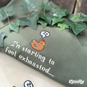 Puede incluir: Sudadera verde salvia con texto bordado en blanco: "I'm starting to feel exhausted..." y un pollo pixelado con una burbuja de pensamiento "zz". El logotipo de Little Big Apparel es visible. Decorada con hojas verdes.