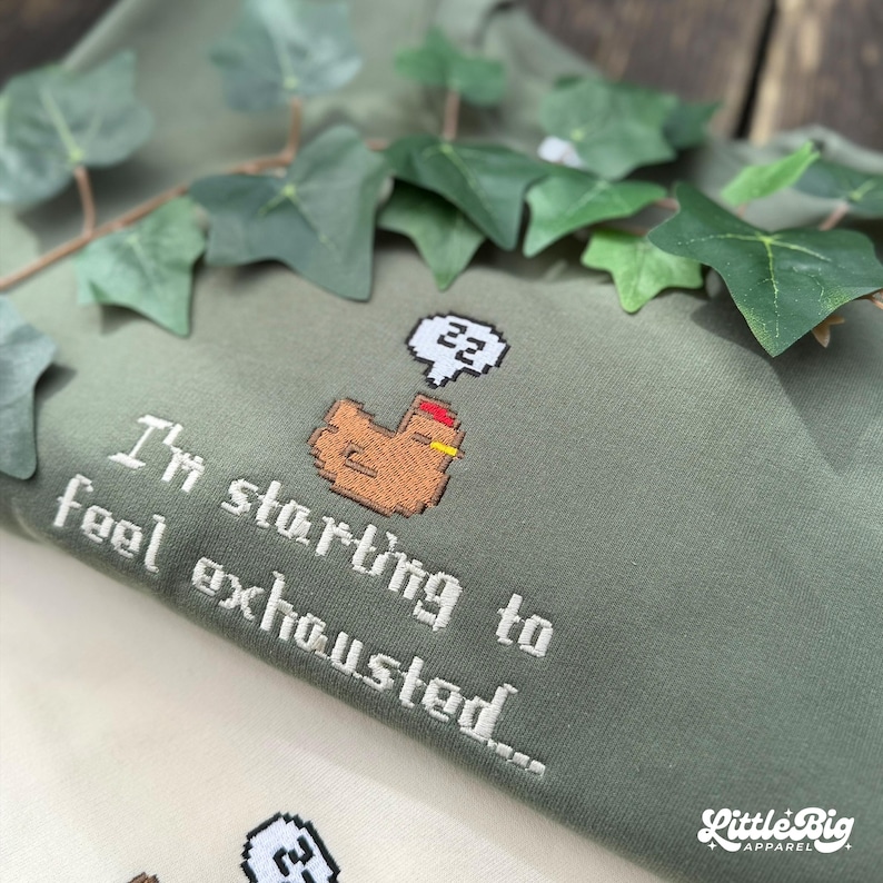 Puede incluir: Sudadera verde salvia con texto bordado en blanco: "I'm starting to feel exhausted..." y un pollo pixelado con una burbuja de pensamiento "zz". El logotipo de Little Big Apparel es visible. Decorada con hojas verdes.