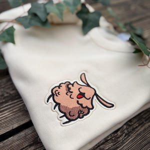 May include: A folded cream-coloured sweatshirt with an embroidered cartoon sheep patch. The sheep has brown fur, a red tongue, and long ears. Green leaves are draped across the top of the sweatshirt.