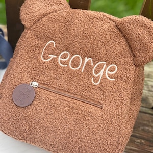 K&ouml;nnte beinhalten: Ein brauner Teddyb&auml;r-Rucksack mit Rei&szlig;verschluss und einem kleinen braunen B&auml;ren-Anh&auml;nger. Der Rucksack ist mit dem Namen "George" in wei&szlig;er Schrift bestickt.