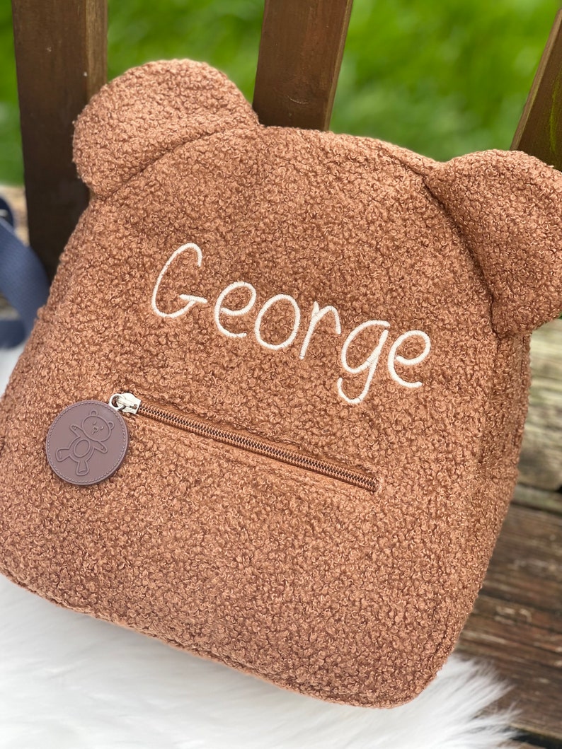 K&ouml;nnte beinhalten: Ein brauner Teddyb&auml;r-Rucksack mit Rei&szlig;verschluss und einem kleinen braunen B&auml;ren-Anh&auml;nger. Der Rucksack ist mit dem Namen "George" in wei&szlig;er Schrift bestickt.