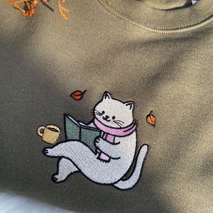 May include: A green sweatshirt with an embroidered design of a white cat reading a book with a pink scarf and a cup of coffee. There are also three small orange leaves embroidered on the sweatshirt.