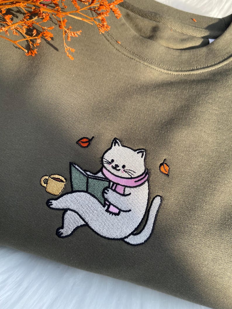 May include: A green sweatshirt with an embroidered design of a white cat reading a book with a pink scarf and a cup of coffee. There are also three small orange leaves embroidered on the sweatshirt.