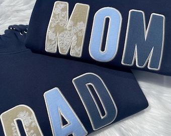 May include: Two navy blue hooded sweatshirts featuring large, block-lettered appliqués. One hoodie displays the word MOM, and the other displays the word DAD. The letters are decorated with a mix of beige floral lace fabric and solid light blue fabric.