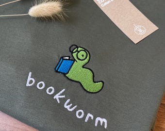 Puede incluir: Una camiseta de color verde oliva con un bordado de un gusano verde claro que lleva gafas y lee un libro azul. Debajo del gusano, la palabra bookworm está bordada en blanco. Tiene una etiqueta marrón con el texto Organic Cotton.