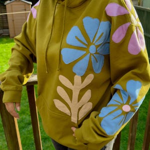 May include: Olive green hoodie with large, appliqued floral designs in light blue, pink, and beige. The hoodie has a kangaroo pocket and a drawstring hood. The floral designs are on the sleeves and the front.