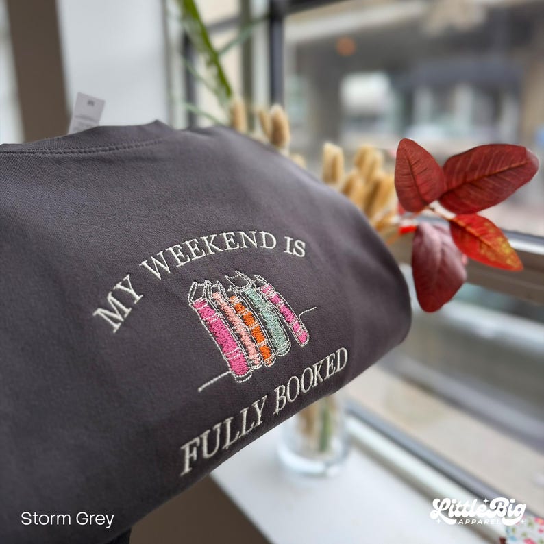 K&ouml;nnte beinhalten: Sturmgraues Sweatshirt mit dem gestickten Text "MY WEEKEND IS FULLY BOOKED" &uuml;ber einer Reihe bunter B&uuml;cher. Das Sweatshirt ist dunkelgrau und hat einen Rundhalsausschnitt.