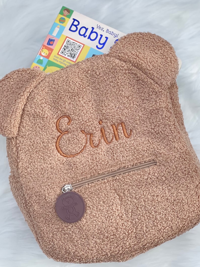 K&ouml;nnte beinhalten: Ein brauner Pl&uuml;sch-Teddyb&auml;r-Rucksack mit einer Rei&szlig;verschlusstasche und einem kleinen braunen B&auml;ren-Anh&auml;nger. Der Rucksack ist mit dem Namen "Erin" in orangefarbenem Garn bestickt.