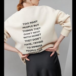 Op de afbeelding: Een crèmekleurig sweatshirt met lange mouwen en zwarte tekst op de achterkant: TOO MANY PEOPLE BUY THINGS THEY DON'T NEED WITH MONEY THEY DON'T HAVE, TRYING TO IMPRESS PEOPLE THEY DON'T LIKE. Het sweatshirt heeft een ronde hals.