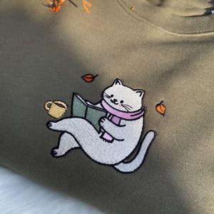 May include: A green t-shirt with an embroidered design of a white cat wearing a pink scarf and reading a book. The cat is holding a cup of coffee and there are falling leaves around it.