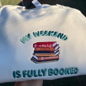 May include: Cream sweatshirt with green embroidered text: "MY WEEKEND IS FULLY BOOKED." A stack of books in red, orange, and brown is embroidered below the text. The sweatshirt is made of a soft material.