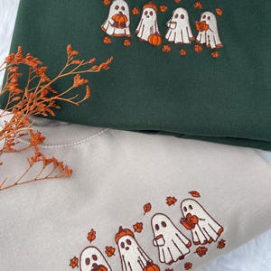 May include: Two sweatshirts, one dark green and one beige, each embroidered with a line of cartoon ghosts holding pumpkins and surrounded by orange leaves. The sweatshirts are folded, with the green one on top.