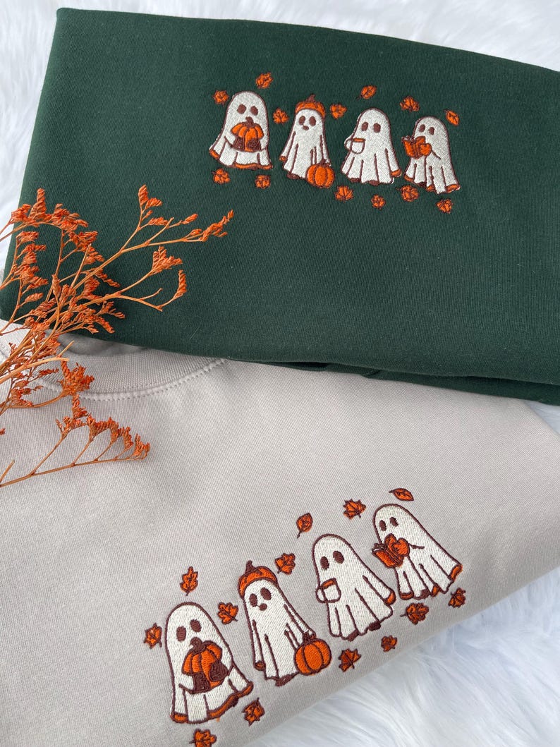 May include: Two sweatshirts, one dark green and one beige, each embroidered with a line of cartoon ghosts holding pumpkins and surrounded by orange leaves. The sweatshirts are folded, with the green one on top.