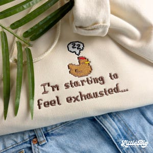 Puede incluir: Sudadera con capucha color crema con cord&oacute;n y un dise&ntilde;o de pollo pixelado. El texto "I'm starting to feel exhausted..." est&aacute; bordado debajo del gr&aacute;fico. La sudadera se combina con unos vaqueros azules.