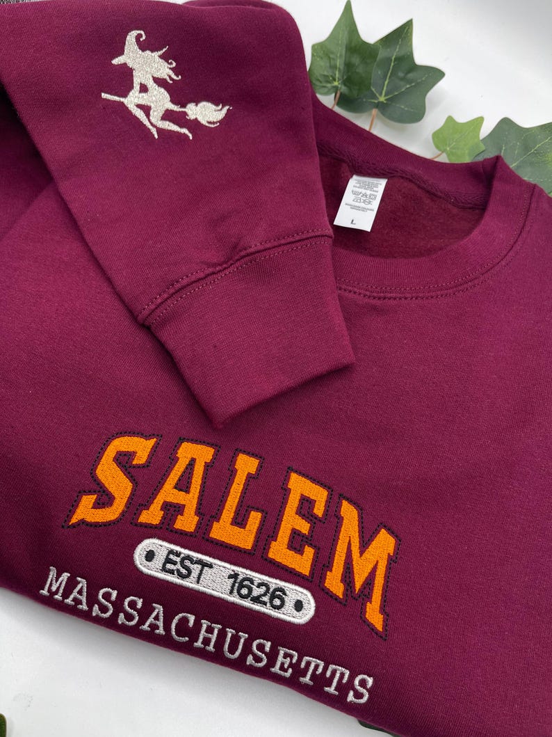 Peut inclure: Sweat-shirt bordeaux avec broderie &laquo; Salem, Massachusetts &raquo; en orange et blanc. La manche pr&eacute;sente une sorci&egrave;re sur un balai. Le texte comprend &laquo; EST 1626 &raquo;.