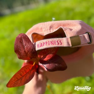 Może przedstawiać: Jasnoróżowa czapka bejsbolowa z napisem "HAPPINESS" wyszytym na różowo na regulowanym pasku. Czapka trzymana jest z gałązką czerwonych liści na rozmytym zielonym tle. Widoczne jest logo Little Big Apparel.