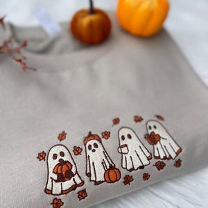 May include: A beige sweatshirt featuring an embroidered design of four cartoon ghosts. Each ghost is holding a pumpkin or a cup of coffee, with autumn leaves scattered around them. Two small orange pumpkins sit on top.