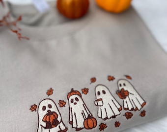 May include: A beige sweatshirt with an embroidered design of four cartoon ghosts. Each ghost is holding a pumpkin or a cup of coffee, with fall leaves scattered around them. Two small orange pumpkins sit on top of the sweatshirt.