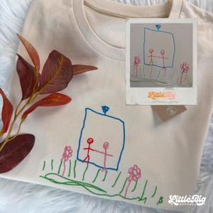 May include: A cream-coloured t-shirt featuring an embroidered design of a blue rectangular frame containing two red and pink stick figures, surrounded by pink flowers and green grass lines. A tag with the text Little Big Apparel is attached.
