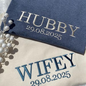 May include: Two t-shirts, one blue and one cream, embroidered with the words "HUBBY" and "WIFEY" in contrasting colors. The date "29.08.2025" is embroidered below each word. The shirts are likely for a couple.