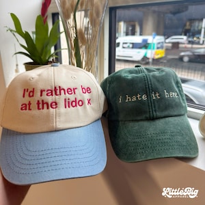 Peut inclure: Une casquette de baseball beige avec une visière bleu clair et un texte brodé en rouge indiquant I'd rather be at the lido x, à côté d'une casquette vert foncé avec un texte brodé en blanc indiquant i hate it here.