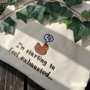 Puede incluir: Sudadera color crema con texto bordado y un dise&ntilde;o de pollo pixelado. El texto dice "I'm starting to feel exhausted..." encima del pollo, que tiene una burbuja de pensamiento con el n&uacute;mero 22.