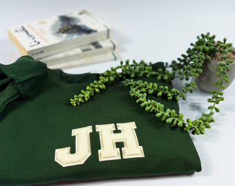 May include: A dark green sweatshirt featuring the embroidered letters "JH" in a cream-coloured, collegiate-style font. The sweatshirt is folded, with a stack of books and a small potted plant with trailing greenery in the background.