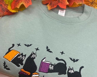 May include: Sage green sweatshirt with an embroidered design of black cats reading books, surrounded by bats and stars. The cats are sitting on stacks of books. Autumn leaves are in the background.