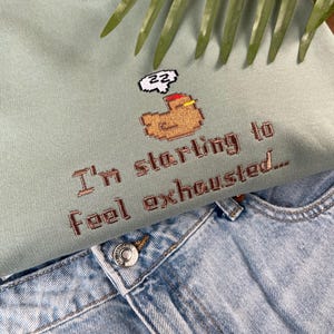 Puede incluir: Una sudadera verde claro con la frase bordada "I'm starting to feel exhausted..." y un gr&aacute;fico pixelado de una figura durmiendo. La sudadera se combina con unos vaqueros de mezclilla azul claro.