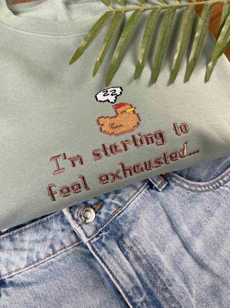 Puede incluir: Una sudadera verde claro con la frase bordada "I'm starting to feel exhausted..." y un gr&aacute;fico pixelado de una figura durmiendo. La sudadera se combina con unos vaqueros de mezclilla azul claro.