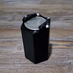 May include: A black hexagonal container with a silver coin inside. The container has a silver top with four small silver screws.