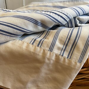 May include: A white and blue striped sheet with a plain white border. The sheet is folded and draped over a wicker basket.