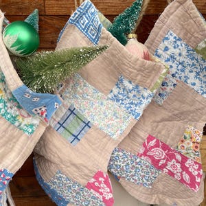 Peut inclure: Chaussettes de Noël faites à la main avec des motifs patchwork dans des tons beige, bleu, rose et vert. Les chaussettes sont décorées de motifs floraux et géométriques. L'image montre également des ornements de Noël verts et roses et de petits arbres décoratifs.