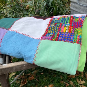 May include: A patchwork quilt featuring a mix of colours and patterns, including green, blue, white, and a floral design. The quilt is folded and resting on a wooden surface.